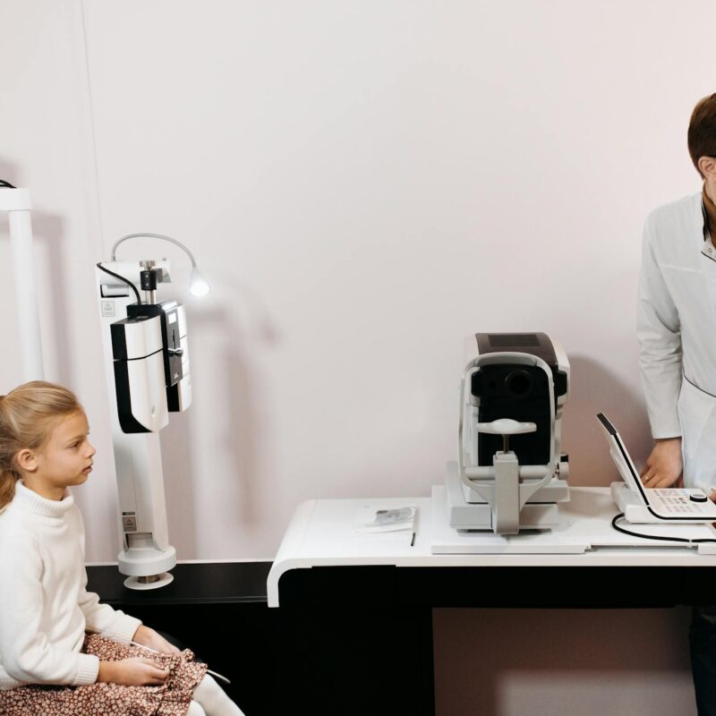 A young girl sits for an eye exam with an optometrist using digital equipment.